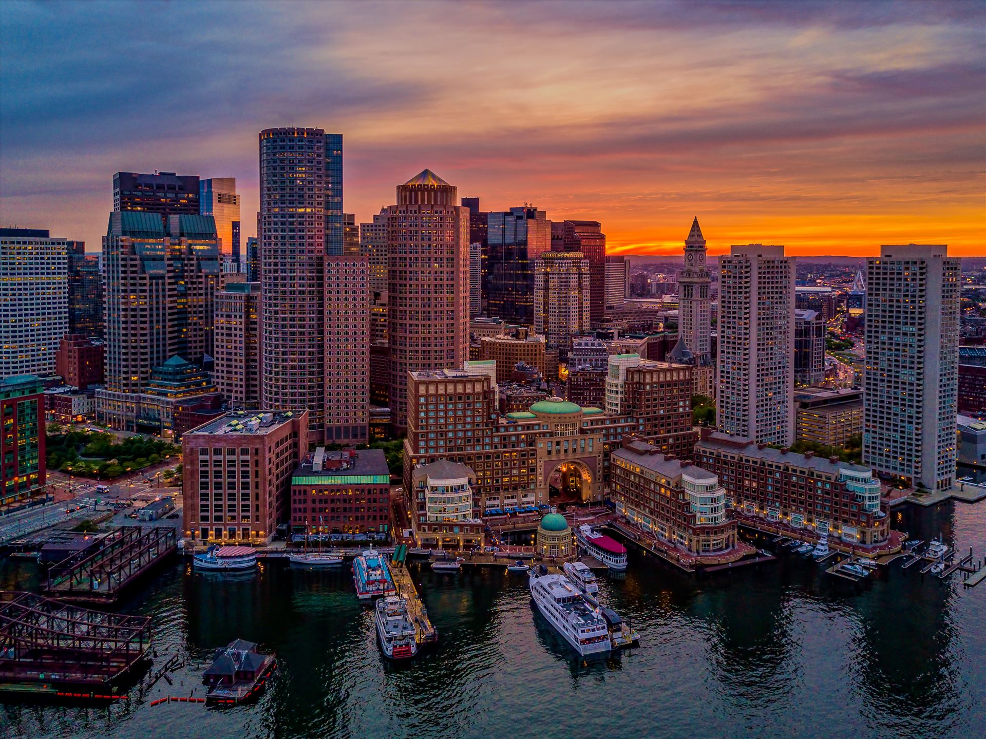 A beautiful sunset illuminates the sky behind the historic downtown Boston waterfront of Boston Harbor in Boston, Massachusetts -  by New England Photography
